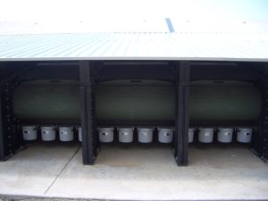 Green cylindrical tank stored under metal roof, with several gray buckets beneath.