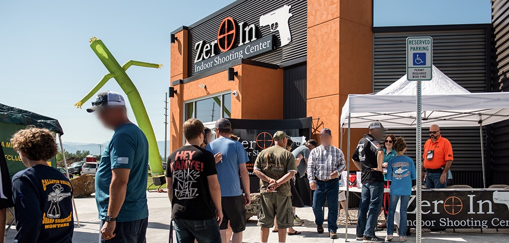 Grand opening event at Zero In Indoor Shooting Center with people gathered outside near tents, signs, and inflatable dancer.