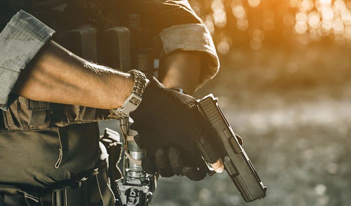Person holding a handgun, dressed in tactical gear, outdoors with sunlight filtering through trees.