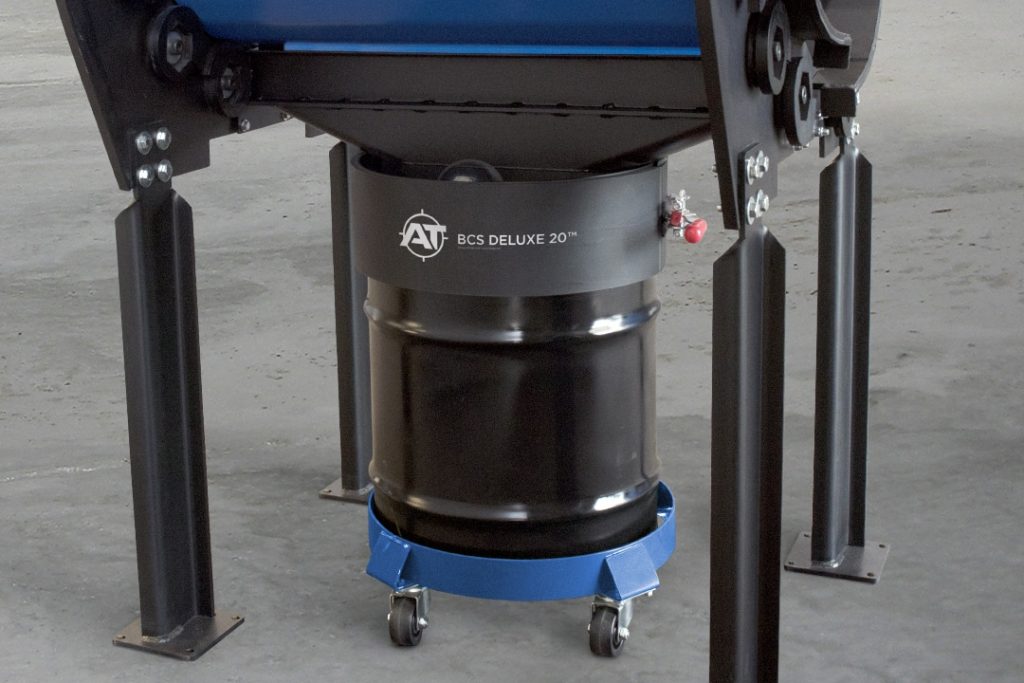 A black drum labeled "AT BCS DELUXE 20™" rests on a wheeled blue base in an industrial setting.