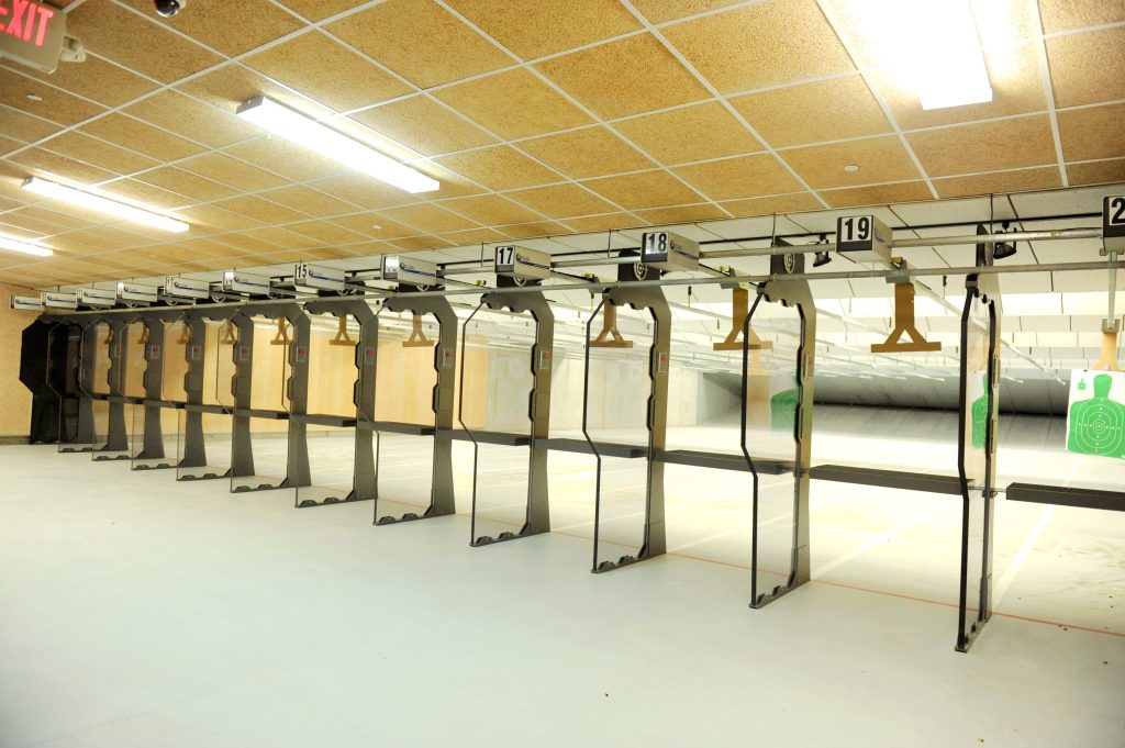 Indoor shooting range with numbered lanes, target holders, and visible exit sign above.