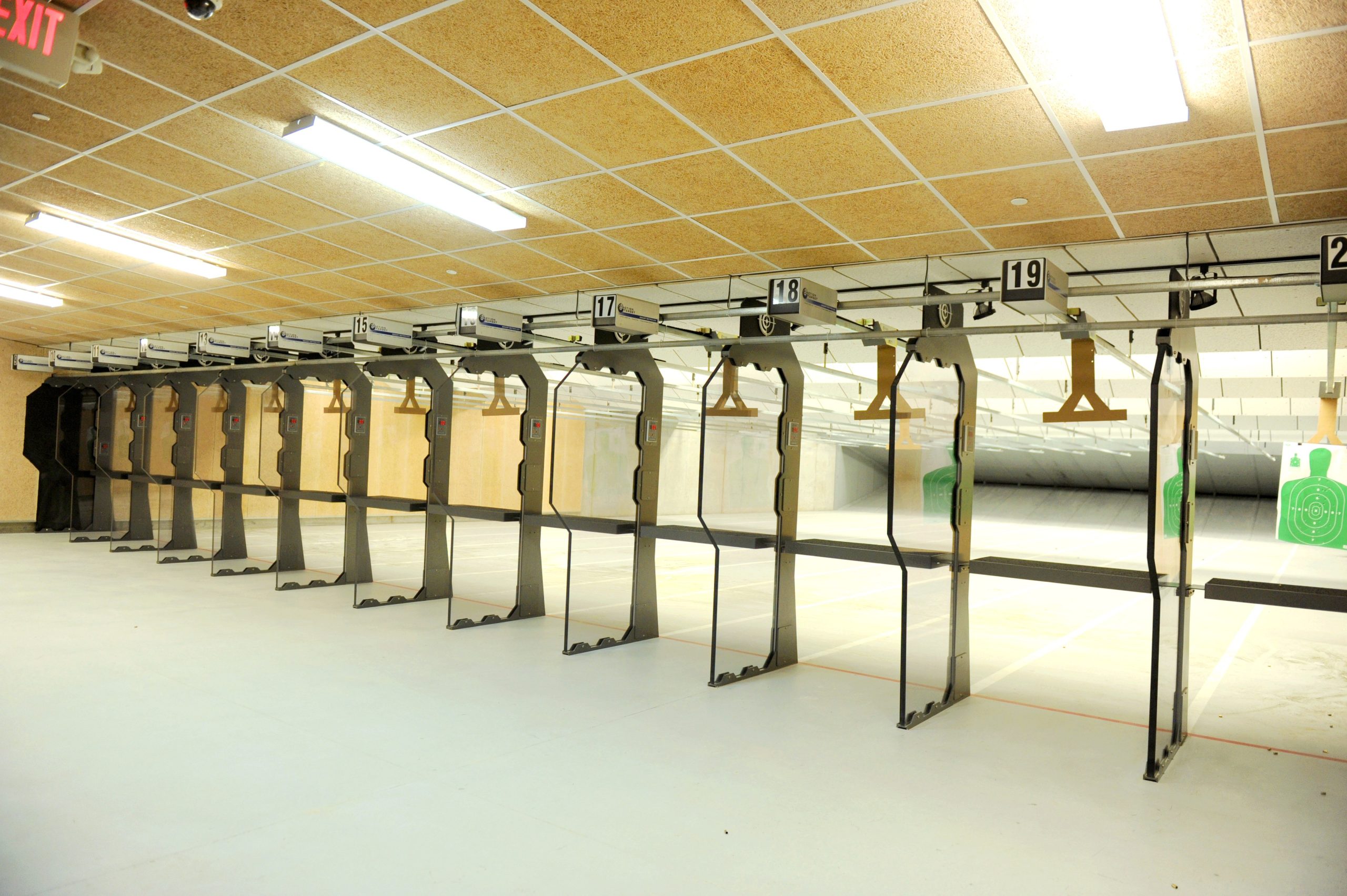 Indoor shooting range with numbered lanes, target holders, and visible exit sign above.