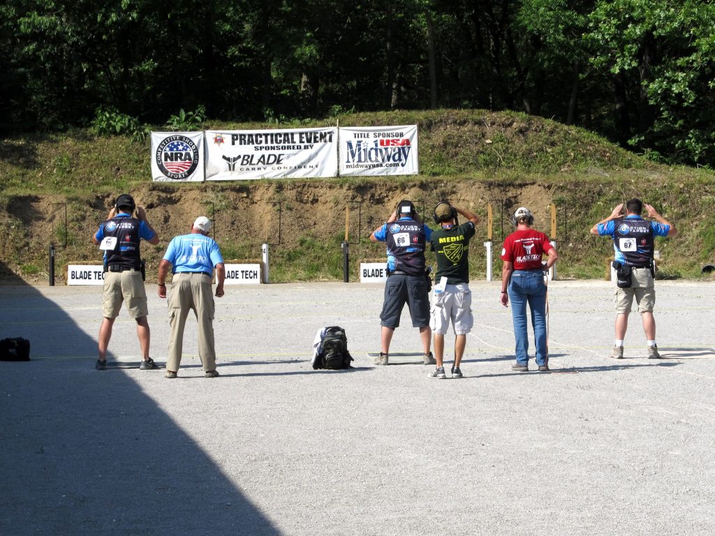 People stand on a shooting range, hands on head, facing targets; banners and trees in the background.