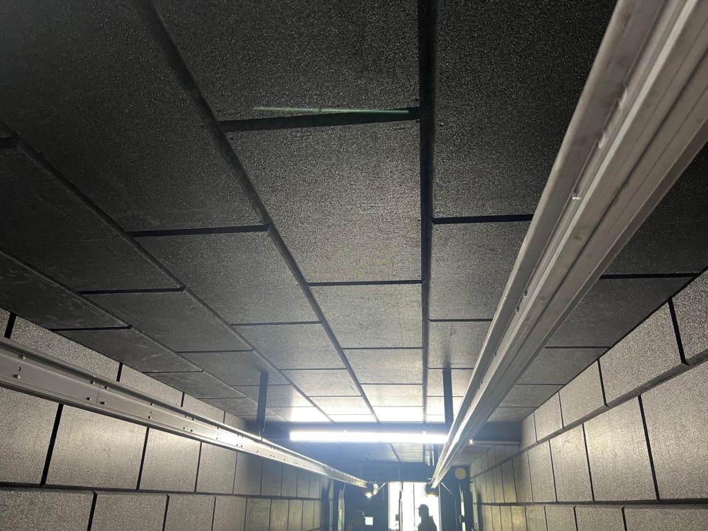 Black ceiling tiles line a tunnel with a lit exit in the distance; silhouette of a person visible.