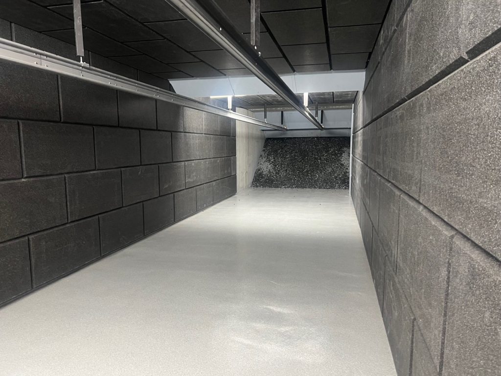 Empty indoor shooting range lane with black acoustic panels, overhead target retrieval system, and bullet trap at the far end.