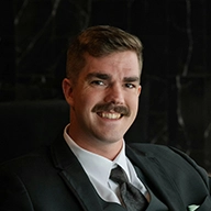 A person in a suit sits smiling in a dark, formal setting, with a black marble background.