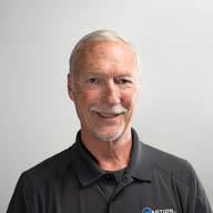 A person smiling, wearing a dark polo shirt, against a plain, light gray background.