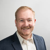 A person smiling, wearing a suit jacket and shirt, against a plain gray background.