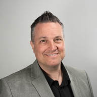 A person smiling, wearing a gray blazer and black shirt, against a plain light gray background.