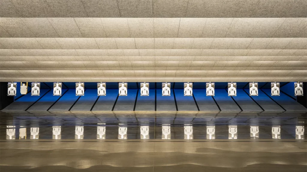 Indoor shooting range with multiple silhouette paper targets aligned in a row under bright ceiling lights.