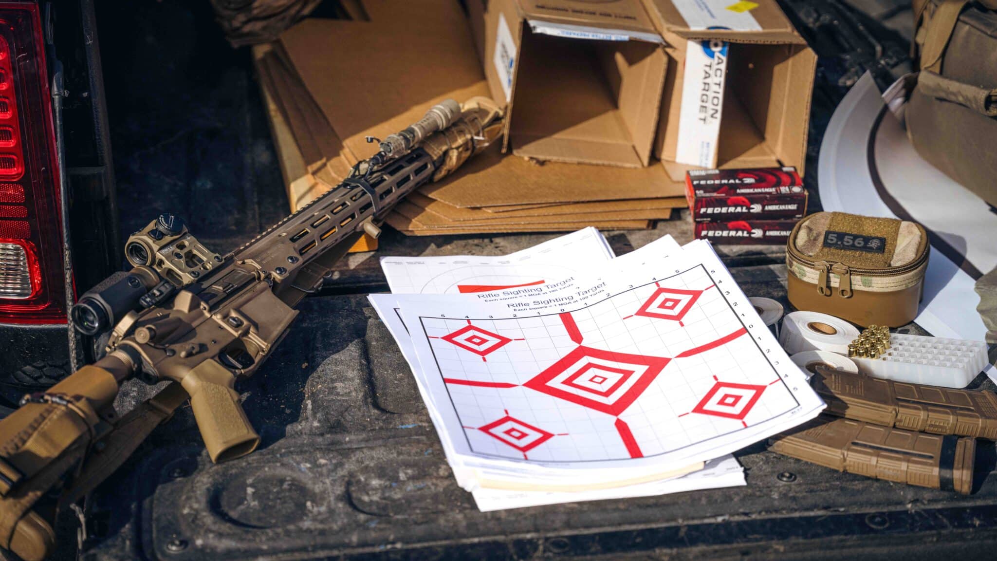 Rifle lying on truck bed with sighting targets, ammo boxes, and cardboard boxes; outdoor setting.