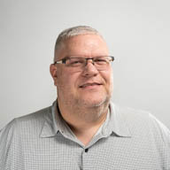 A person with glasses smiles slightly, wearing a gray shirt, against a plain background.