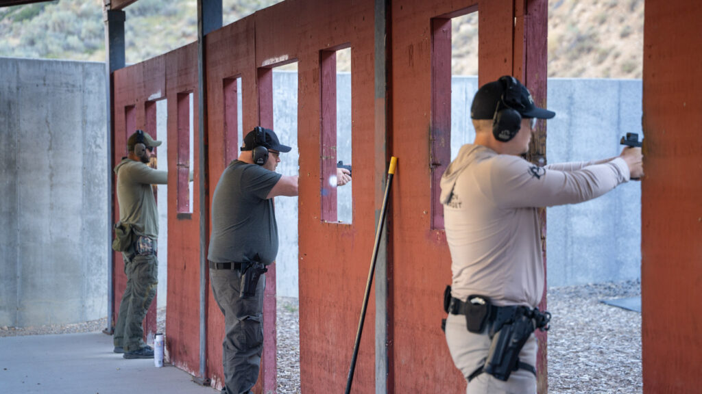 Three people aim pistols at targets in a shooting range, divided by red partitions.