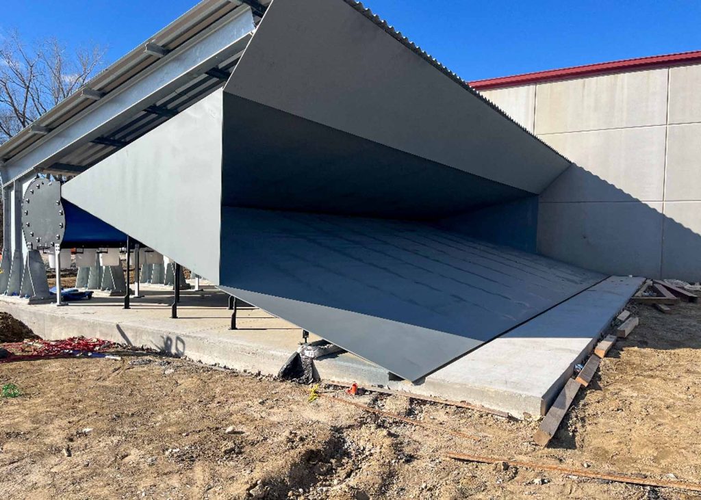 Large gray chute, angled upward, next to industrial structure on bare ground.