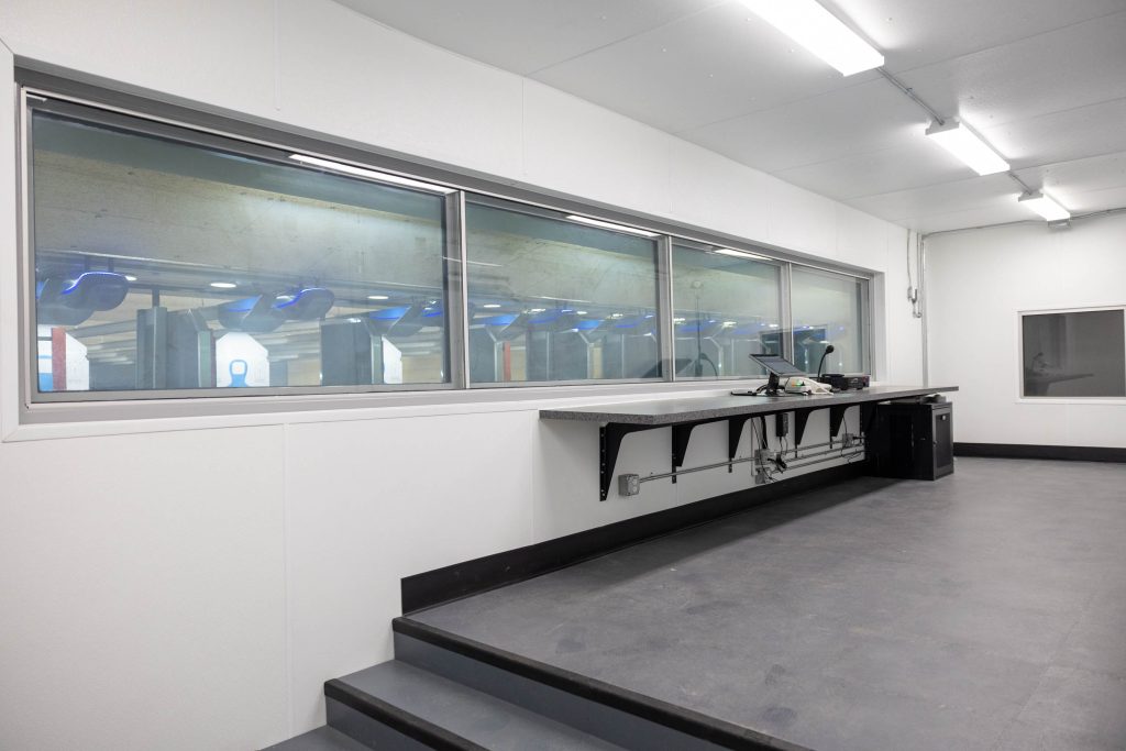 Room with a long window overlooking a shooting range; counter with a computer in a bright space.